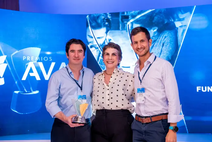Dos hombres en escenario con una mujer, uno de ellos sostiene un premio AVA